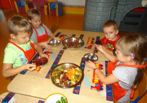 Dzieci siedzą przy stole. Chłopiec i dziewczynka trzymają w ręku łyżki którymi przekładają maliny i borówki do dużej miski.